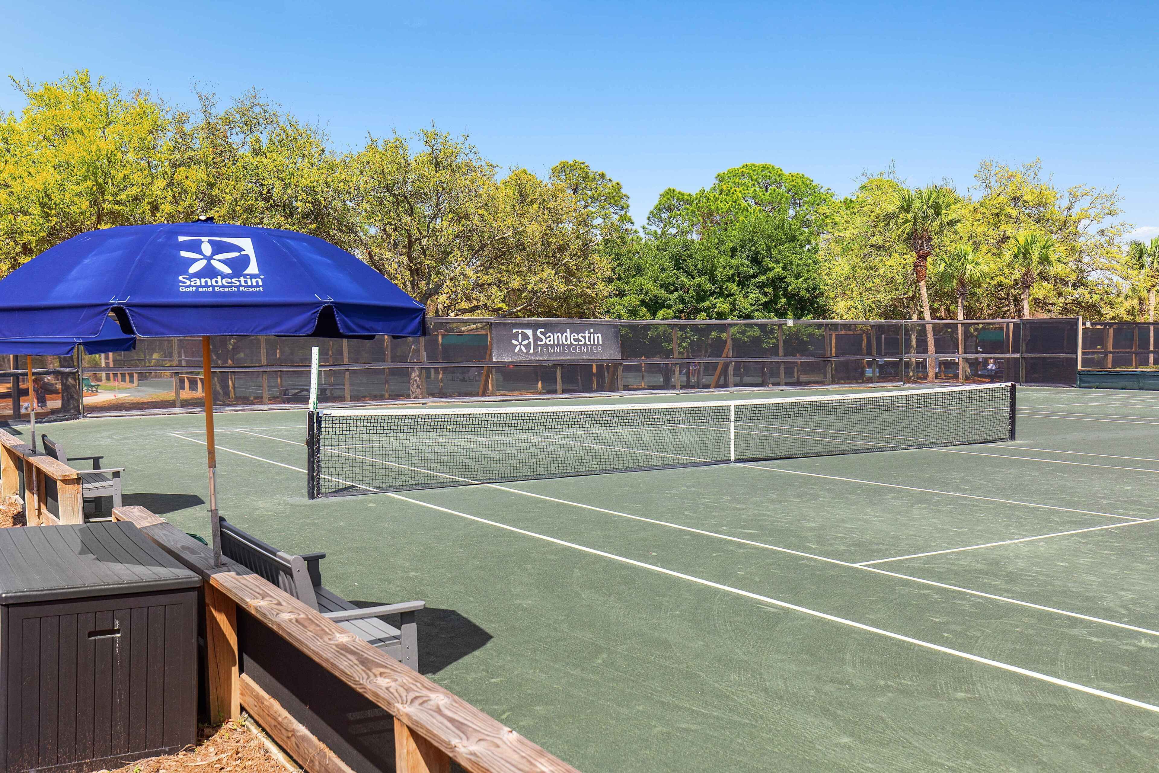 A tennis court with a net, wooden benches, and a blue umbrella.