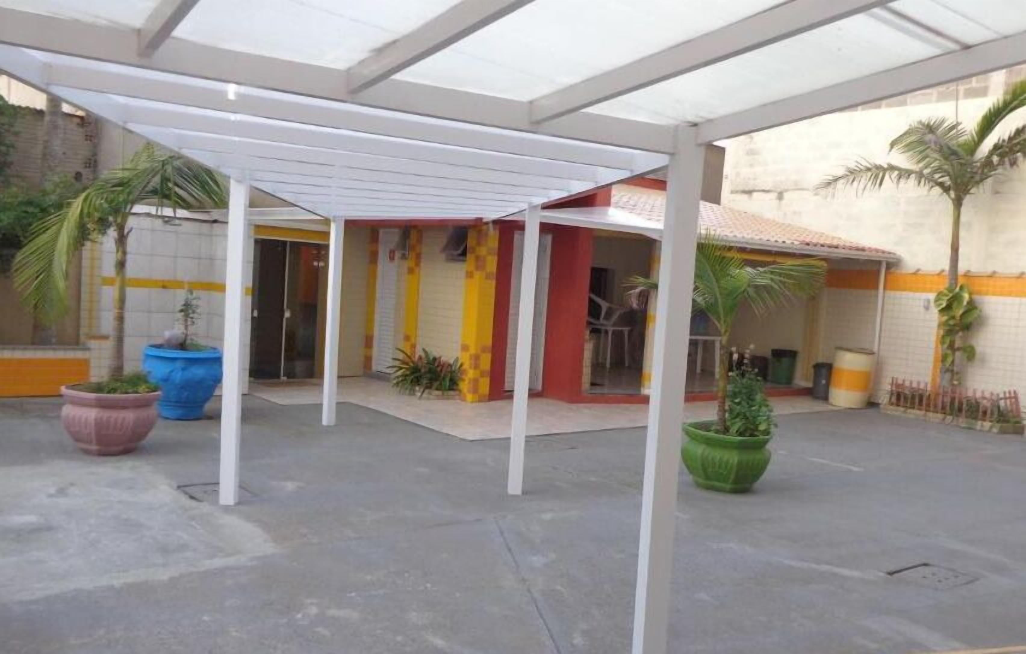 A covered outdoor area with a white pergola, potted plants, and a building entrance with a tiled wall.