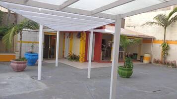 A covered outdoor area with a white pergola, potted plants, and a building entrance with a tiled wall.