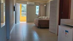 A spacious room with a bed, a small table, and a refrigerator. There is a sliding door leading to a balcony. - Lebanon Praia Hotel (Tramandai)