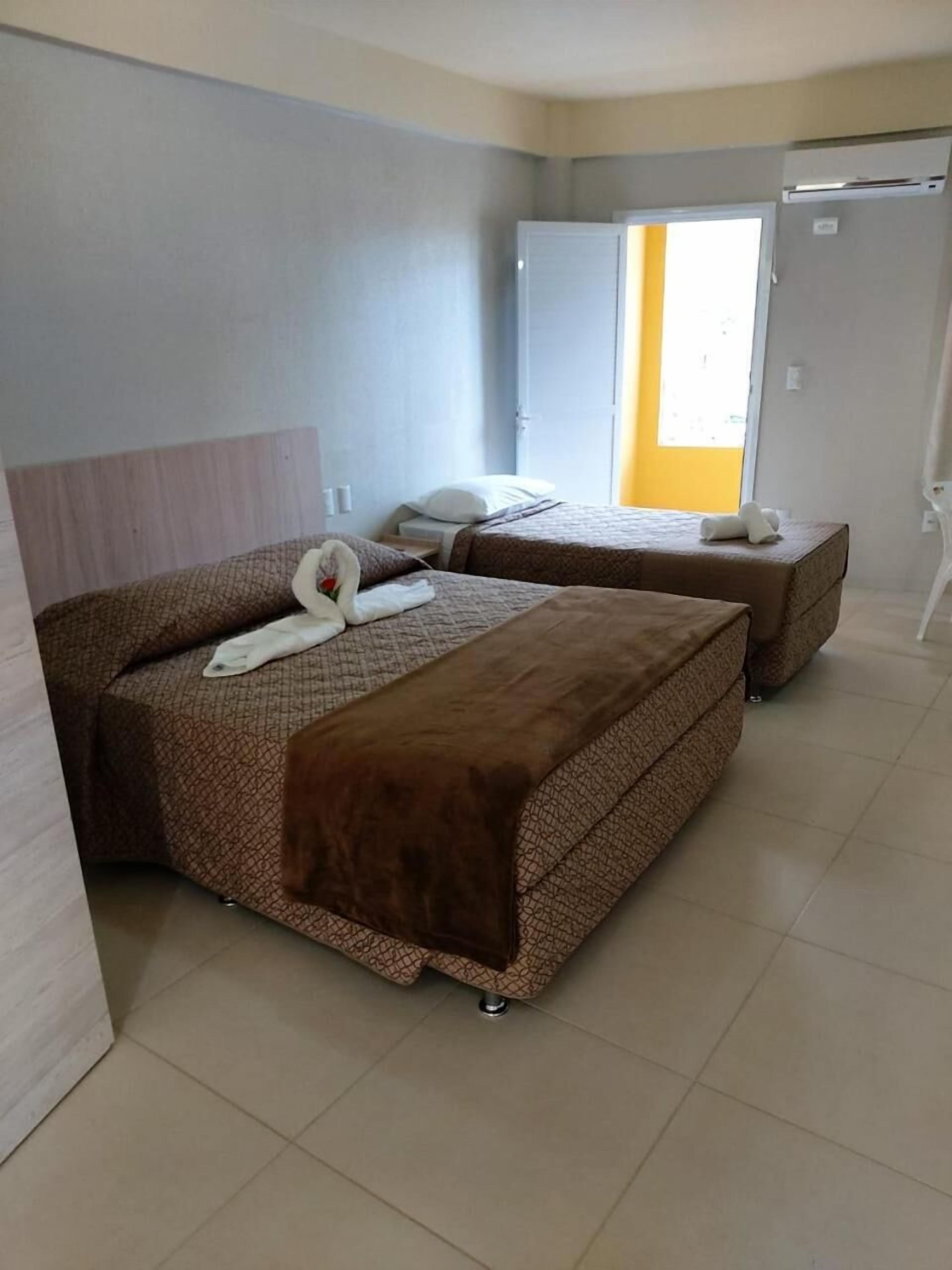 A hotel room with two beds, a wooden headboard, a white wall, and a door leading to a bright room.