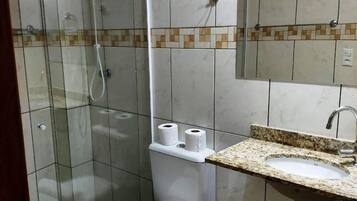 Casa de banho com chuveiro em cabine de vidro, sanita, lavatório com espelho e um contentor de lixo.