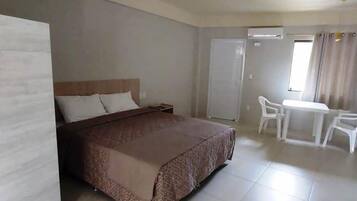 Habitación de hotel con una cama grande, una pequeña mesa con sillas, un equipo de aire acondicionado y un ventanal con cortinas.
