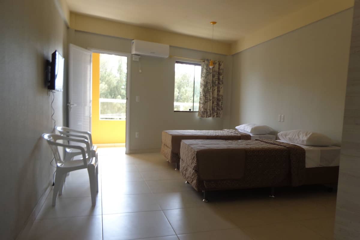A room with two beds, a chair, a window with curtains, and an air conditioning unit.