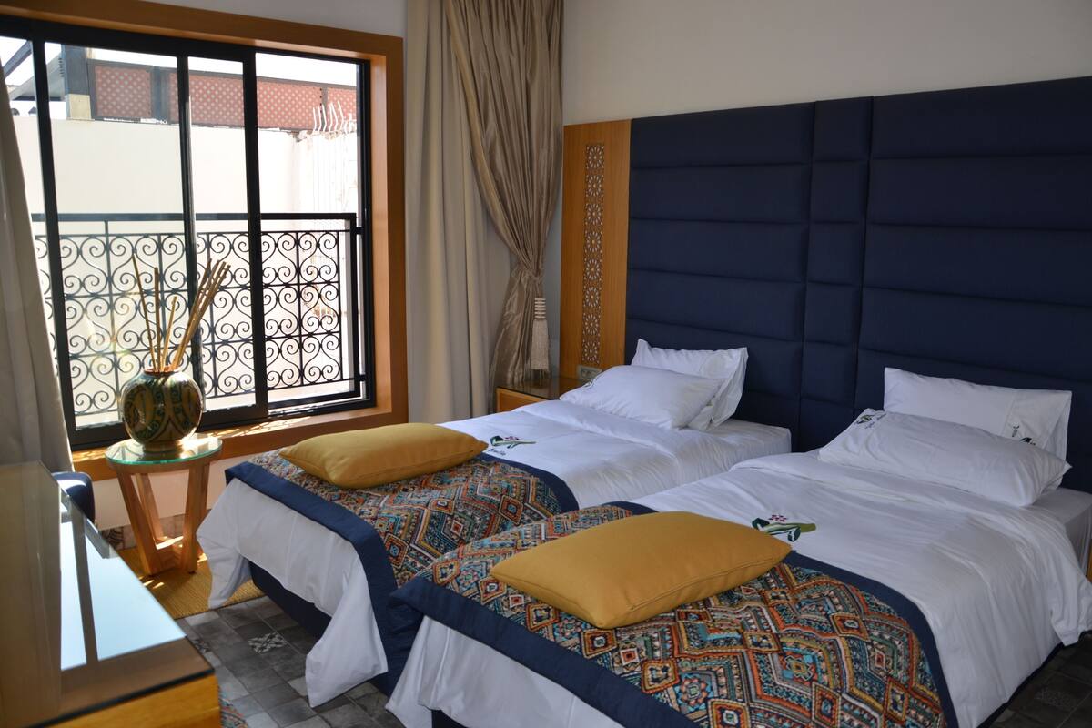 Ein Hotelzimmer mit zwei Betten, einem großen Fenster mit Blick, einem kleinen Tisch mit einer Vase und einem hölzernen Kopfteil.
