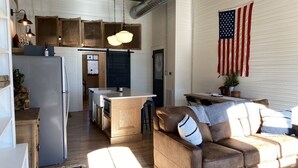 Una cocina compacta con refrigerador, gabinetes de madera y una isla central. En el área de estar, un sofá de cuero marrón y una mesa de centro, con una bandera estadounidense en la pared.