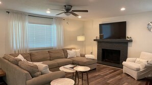 Un salon comprenant un canapé d’angle, une cheminée, un ventilateur de plafond et un t éléviseur fixé au mur au-dessus de la cheminée.