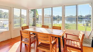 A sunlit room with a large table and chairs, large windows with a view of a lake and greenery.