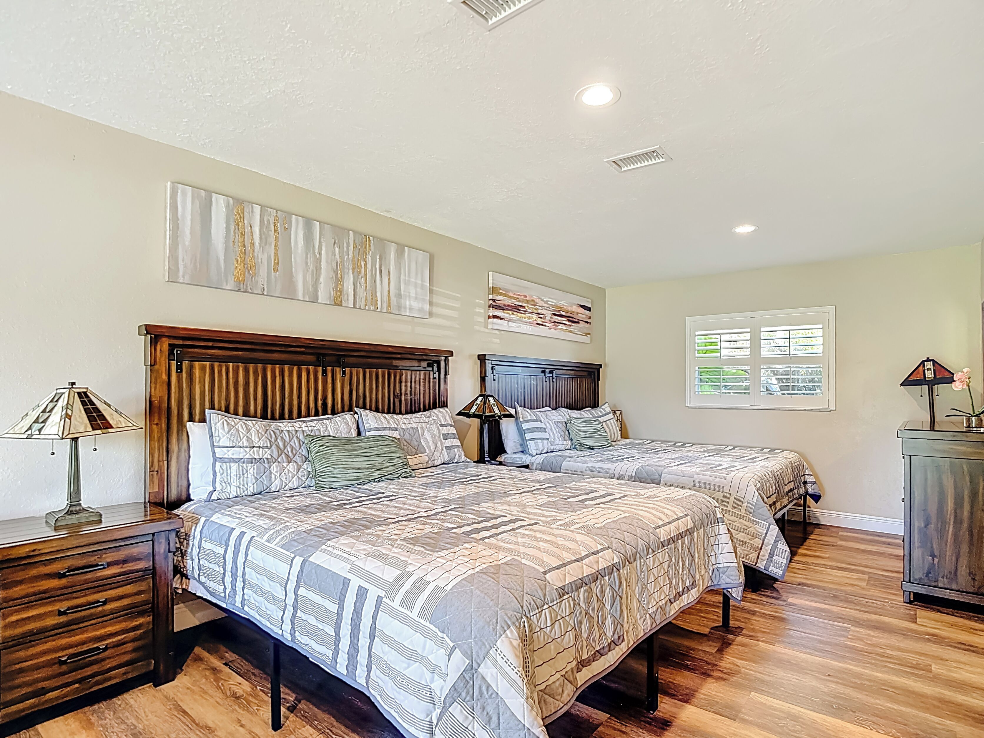 A bedroom with two beds, wooden headboards, a nightstand with a lamp, and artwork on the walls.