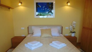 Ein ordentlich bezogenes Bett mit einer beigen Steudecke, zwei Kissen und zwei gefalteten Handtüchern. An der Wand über dem Bett hängt ein gerahmtes Bild mit einer Strandansicht.