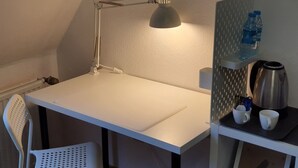 Un petit bureau blanc, surmonté d’une seule lampe, avec une chaise blanche, un radiateur, une bouteille d’eau et une bouilloire posée sur une étagère.