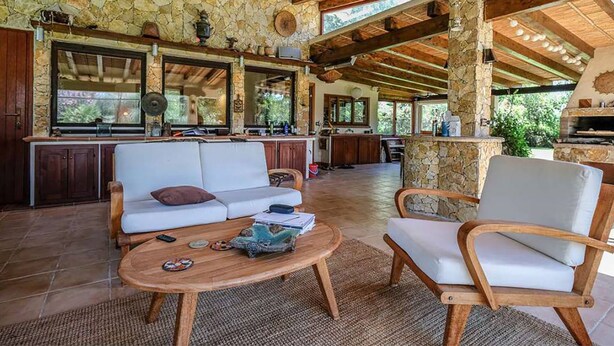 A spacious living area with a stone wall, wooden ceiling beams, a sofa, chairs, and a round coffee table. - Casa Rustico, large pool, 350m from the beach, by the river. (Chia)