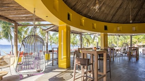 Un restaurant situé en bord de mer, avec une façade jaune incurvée, des tables et des chaises en bois, et un toit de chaume.