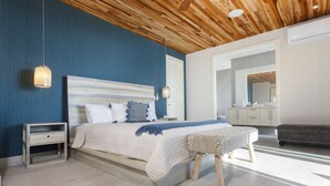 Un dormitorio con techo de madera, una cama grande con cabecera azul y blanca, mesitas de noche, un banco y un espejo grande.