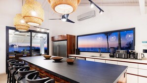 A modern kitchen with a large island, bar stools, and a view of the sunset through a large window. - Beachfront Cliff Villa • 300° Ocean Views • Pool, Jacuzzi & Guest House (Playa Flamingo)