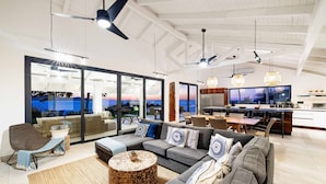 A spacious living area with a large sofa, a coffee table, and a television. There is a kitchen area with a dining table and chairs. The room has large windows with a view of the outdoors. - Beachfront Cliff Villa • 300° Ocean Views • Pool, Jacuzzi & Guest House (Playa Flamingo)