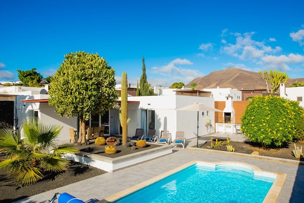 A pool area with a clear blue pool, surrounded by white buildings, cacti, and a mountain in the background.