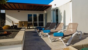 A sunlit outdoor seating area with lounge chairs and a table, surrounded by a low stone wall and a wooden pergola.