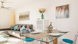 A modern living room with a glass dining table, clear chairs, a sofa with cushions, a painting on the wall, and a vase with flowers.