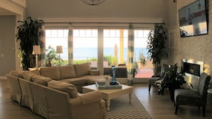 A living room with a sectional sofa, a coffee table, and a TV on the wall. There are potted plants and a view of the ocean through large windows. - Southampton  PRIVATE Waterfront Estate!
 (Hampton Bays)