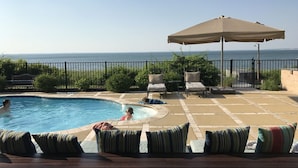 A swimming pool with lounge chairs and an umbrella, surrounded by a fence and greenery, overlooking a body of water. - Southampton  PRIVATE Waterfront Estate!
 (Hampton Bays)