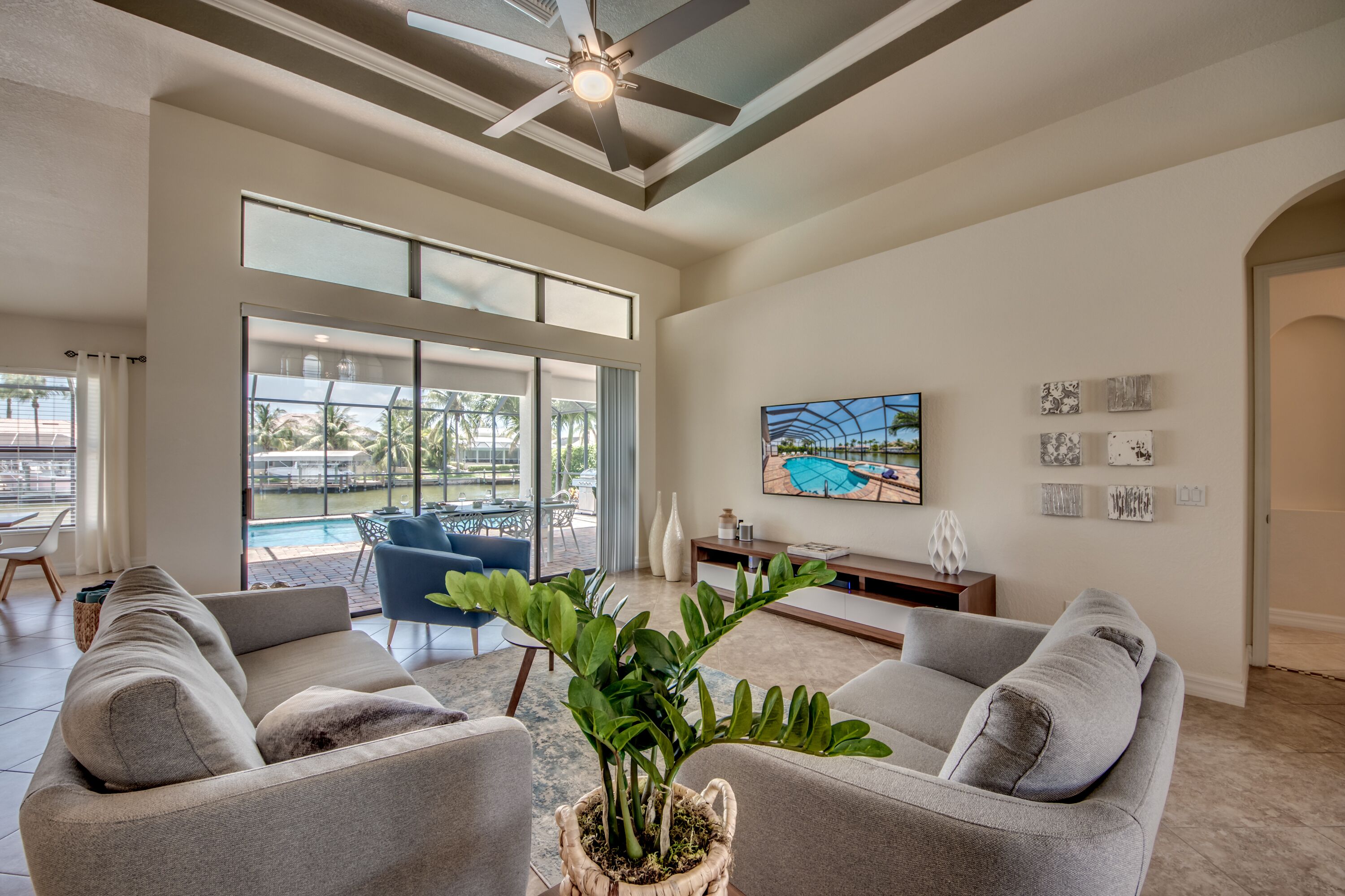 A spacious living room with a grey sofa set, a large window overlooking a pool, and a ceiling fan.