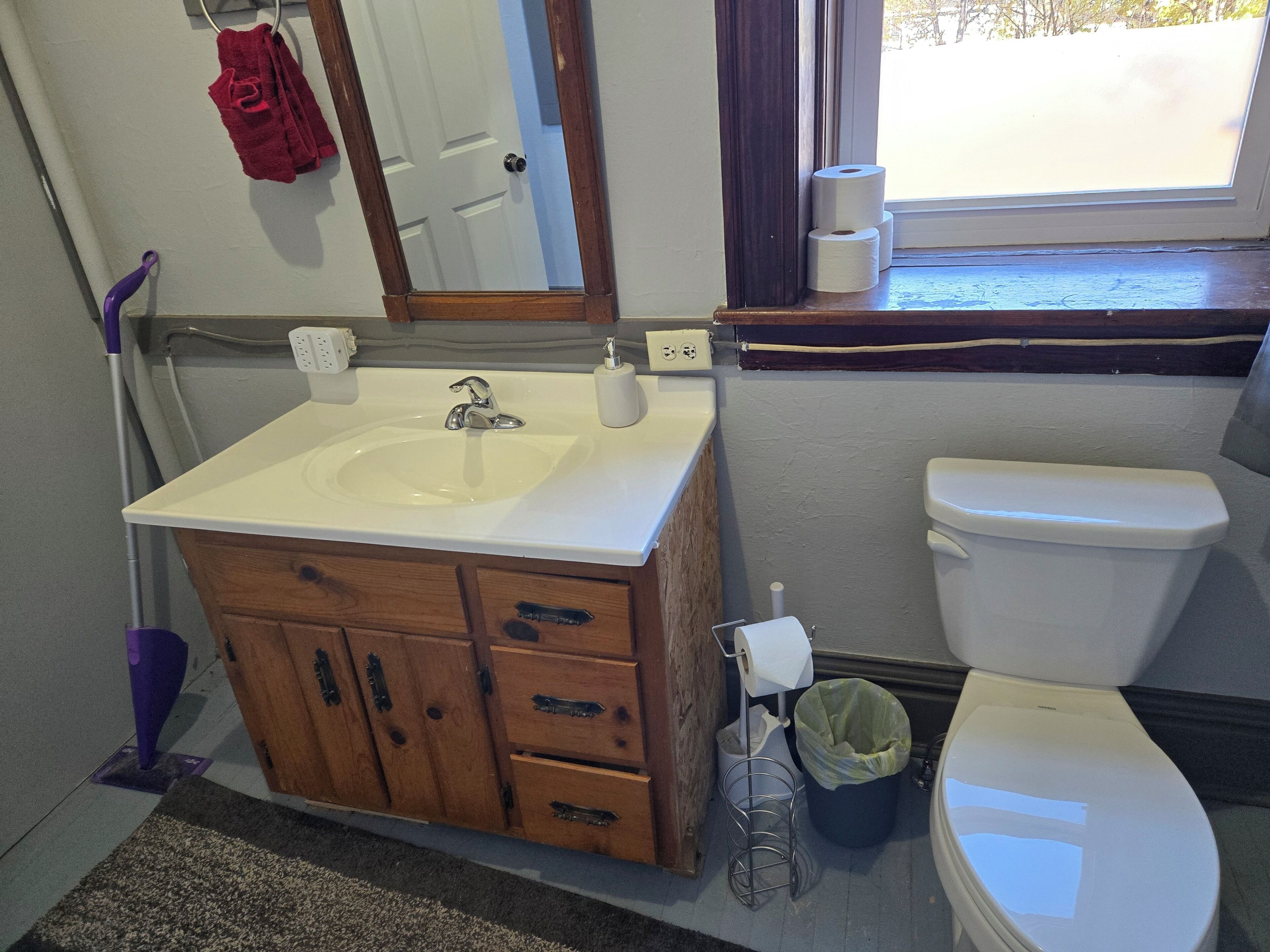 Een badkamer met een houten wastafelmeubel, een wit wastafel, een spiegel, een toilet en een raam met uitzicht op bomen.