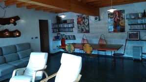 A cozy living room with a wooden ceiling, a grey sofa, a dining table with chairs, and a bookshelf filled with books. - Hobbit House (bioclimatic and half-buried with green cover). (Alba de los Cardaños)