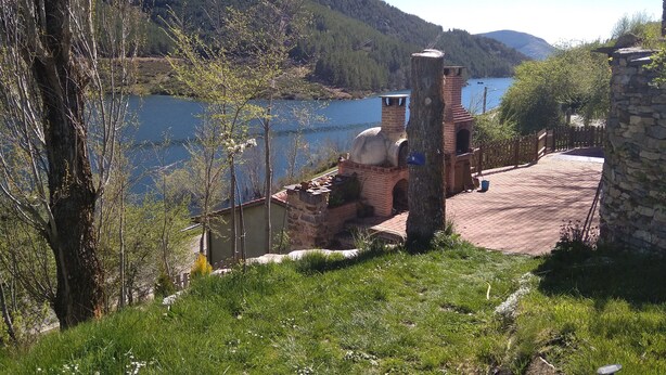 A rustic building with a brick chimney and a stone tower, surrounded by lush greenery and a lake. - Hobbit House (bioclimatic and half-buried with green cover). (Alba de los Cardaños)