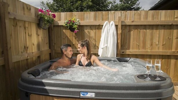A wooden deck with a hot tub, two people in the tub, and two glasses of champagne on the side.