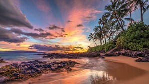 Ein Strand mit Sonnenuntergang, Palmen und felsiger Küste.