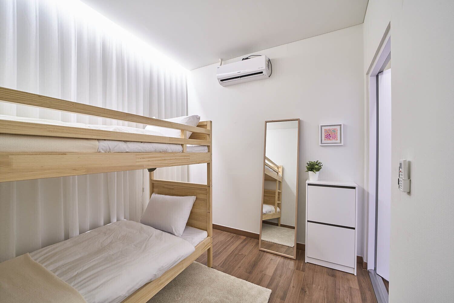 Uma cama beliche com estrutura de madeira e roupa de cama branca, um pequeno armário branco, um espelho e um aparelho de ar condicionado na parede.