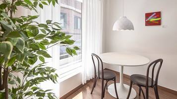 Um quarto com uma mesa redonda e quatro cadeiras, uma luminária pendente, um vaso de planta e uma janela com cortinas.
