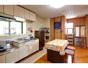 Eine kleine Küche mit Mikrowelle, Kühlschrank und Holzausstattung. Ein Tisch mit Blumenmuster bedeckt, und zwei Hocker stehen daneben. Eine Dartscheibe ist an der Wand angebracht.