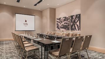 Un salón de conferencias con una larga mesa, sillas, una pantalla de proyector y un cuadro abstracto grande en la pared.
