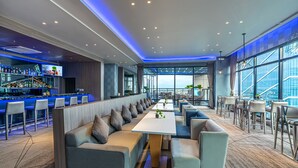 A modern bar area with a long counter, seating with cushions, and a view of the city through large windows.