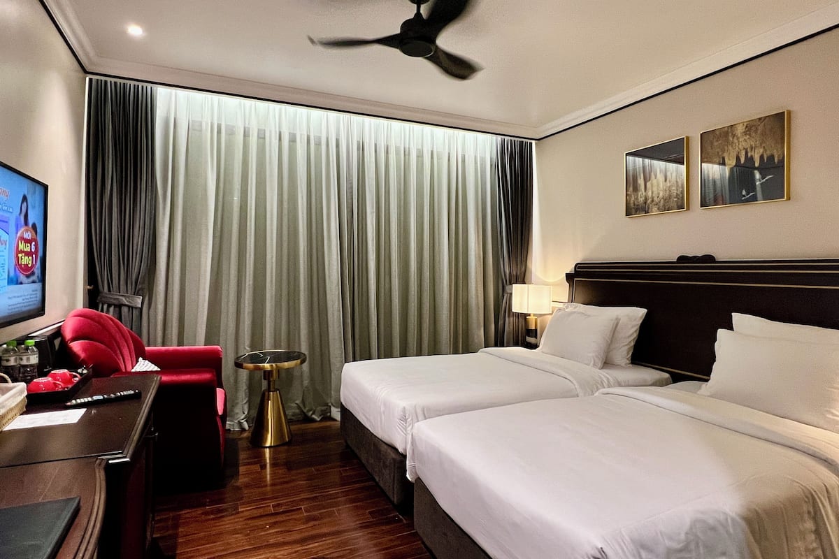 A hotel room with two beds, a desk with a chair, a TV, a ceiling fan, and a window with curtains.