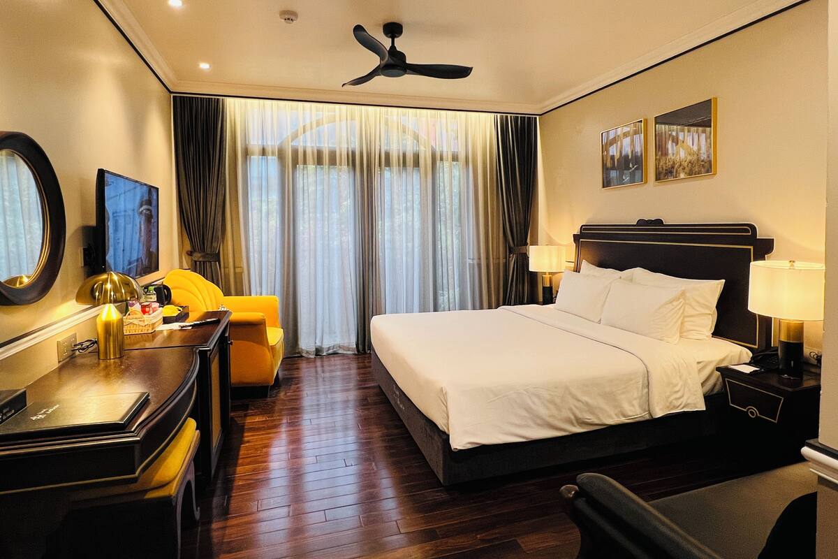A hotel room with a large bed, a desk with a chair, a desk lamp, a television, and a ceiling fan.