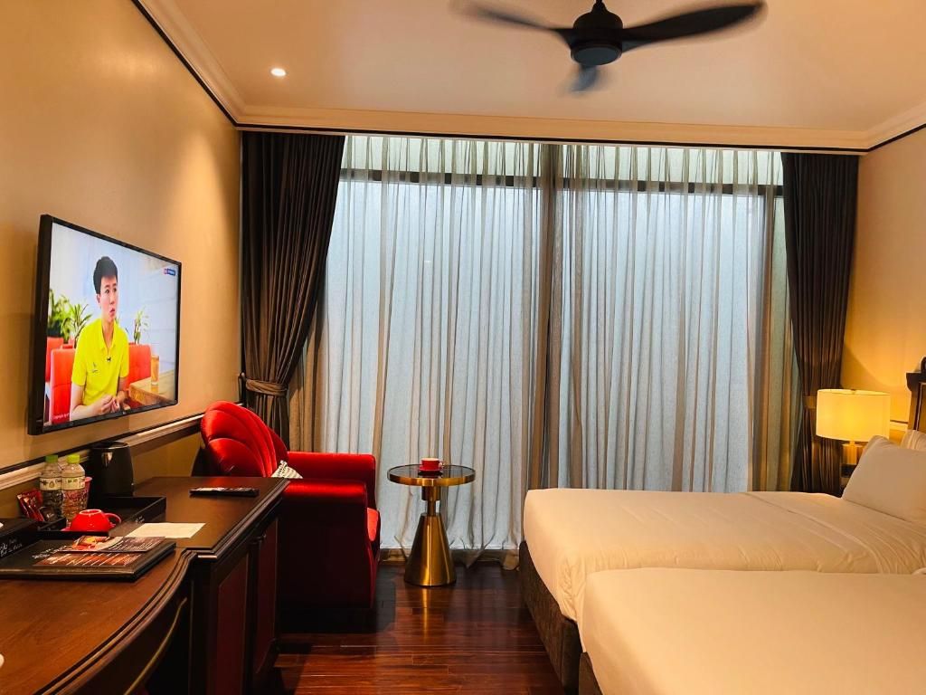 A hotel room with a bed, a desk with a television, a red chair, a small round table, and a ceiling fan.