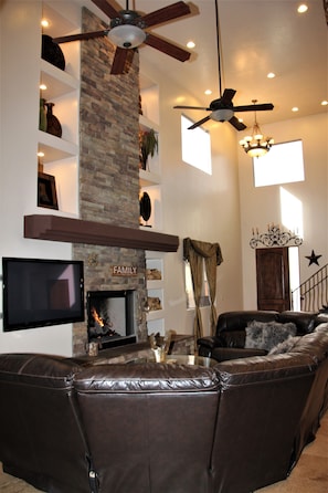 A living room with a fireplace, leather sofa, ceiling fans, and a TV.