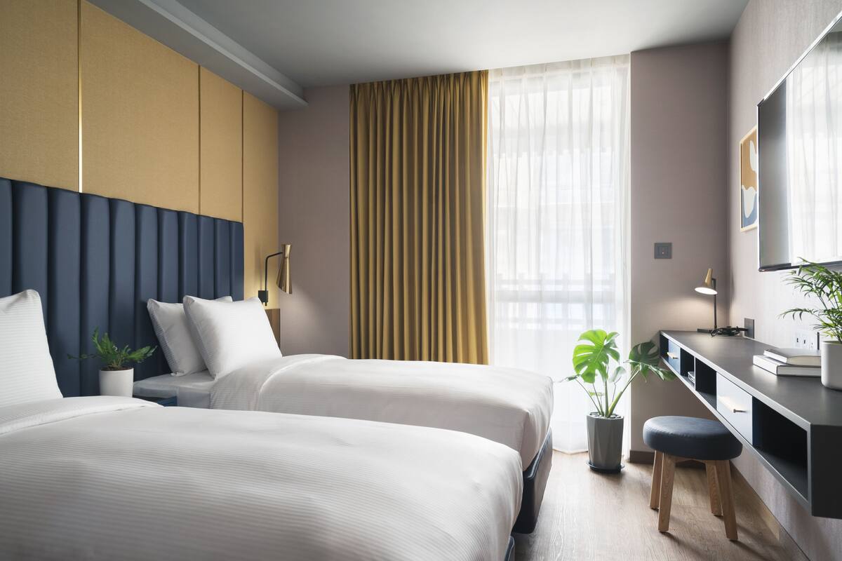 A hotel room with two beds, a desk, a chair, a potted plant, and a large window with curtains.