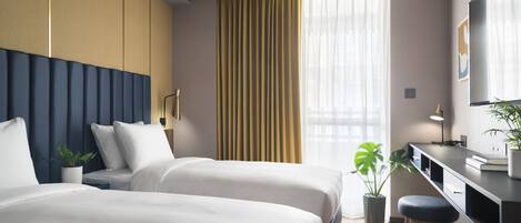 Habitación de hotel con dos camas, un escritorio, una silla, una planta en maceta y un ventanal con cortinas.