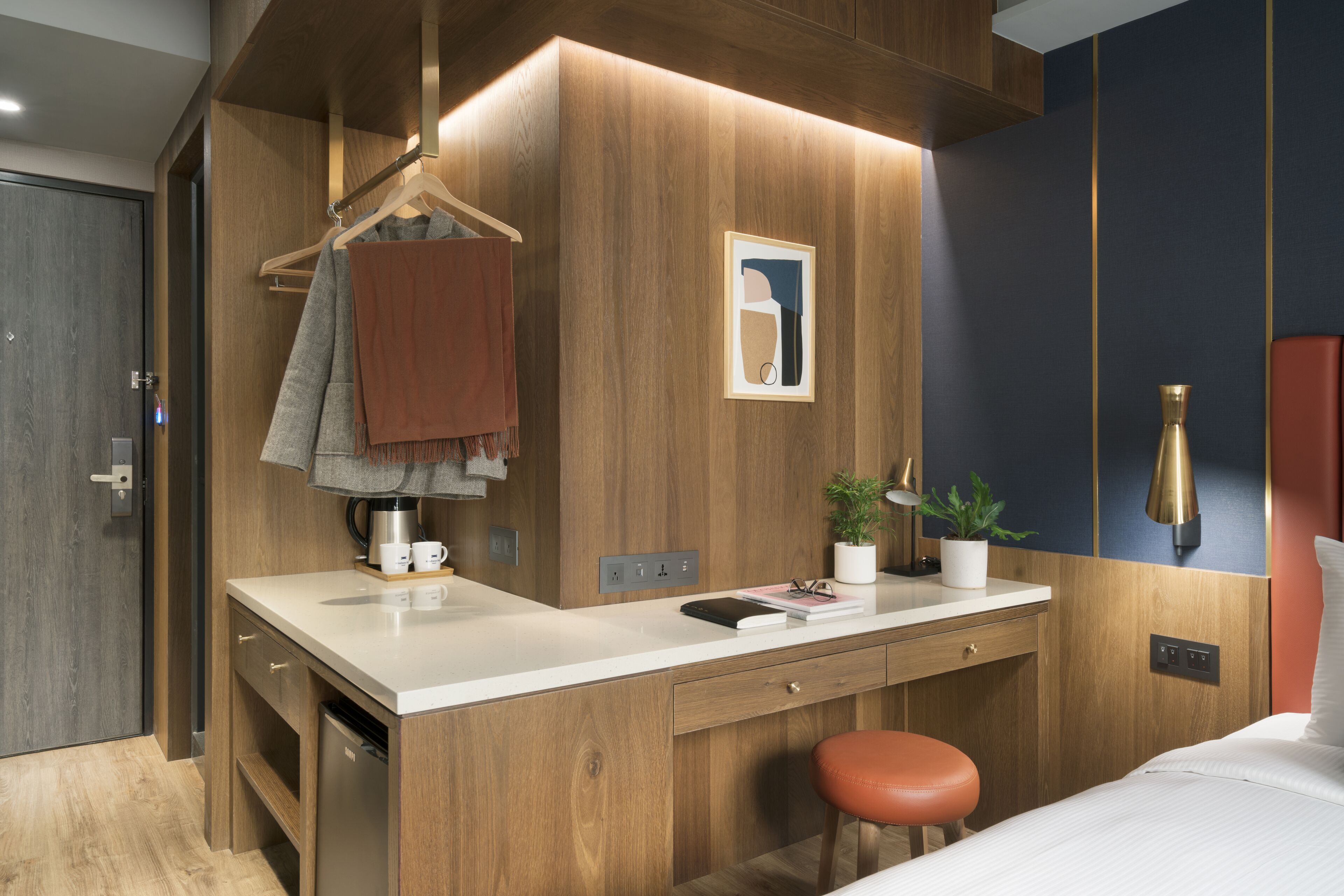 A modern hotel room with a wooden accent wall, a desk with a coffee machine, a hanging towel, and a bed with a red cushion.