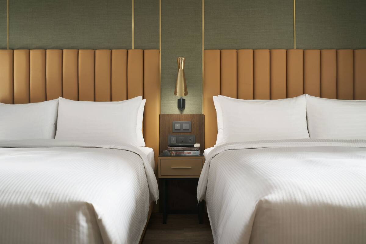 A hotel room with two beds, a nightstand with a lamp, and a wall-mounted control panel.