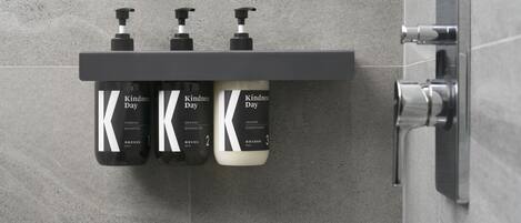 Un baño con pared de azulejos grises y una repisa con tres botellas de productos Kindness Day.