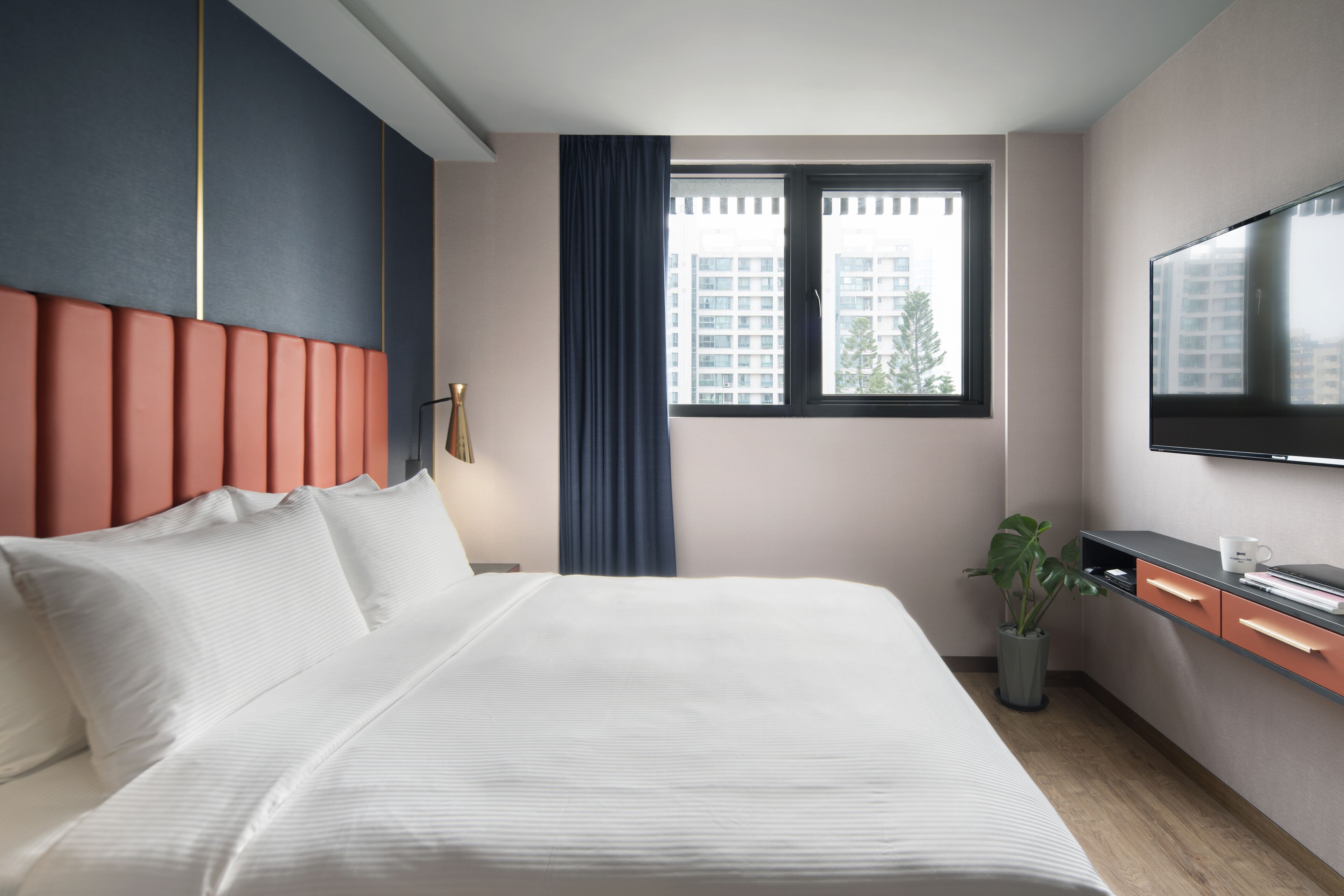 Habitación de hotel con cama, escritorio con televisor de pantalla plana, ventana con cortinas y una planta en maceta.