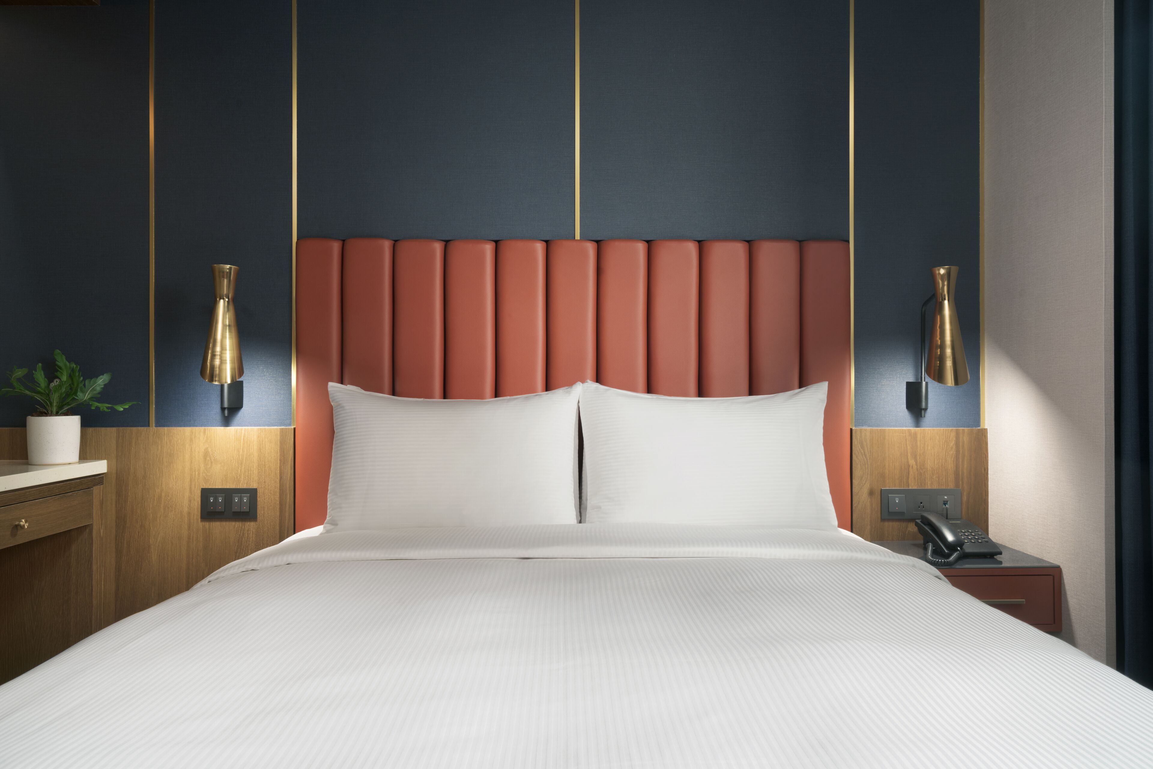 A hotel room with a large bed, wooden headboard, two bedside tables with lamps, a small plant, and a telephone.