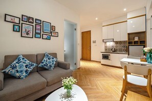 A modern living room with a sofa, coffee table, and wall art. A kitchen with white cabinets and a dining area with a table and chairs is visible in the background.