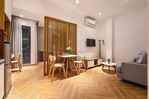 A modern living room with a herringbone wooden floor, a grey sofa, a dining table with chairs, and a TV mounted on the wall. - The Art - Sunshine Apartment (Ho Chi Minh City)