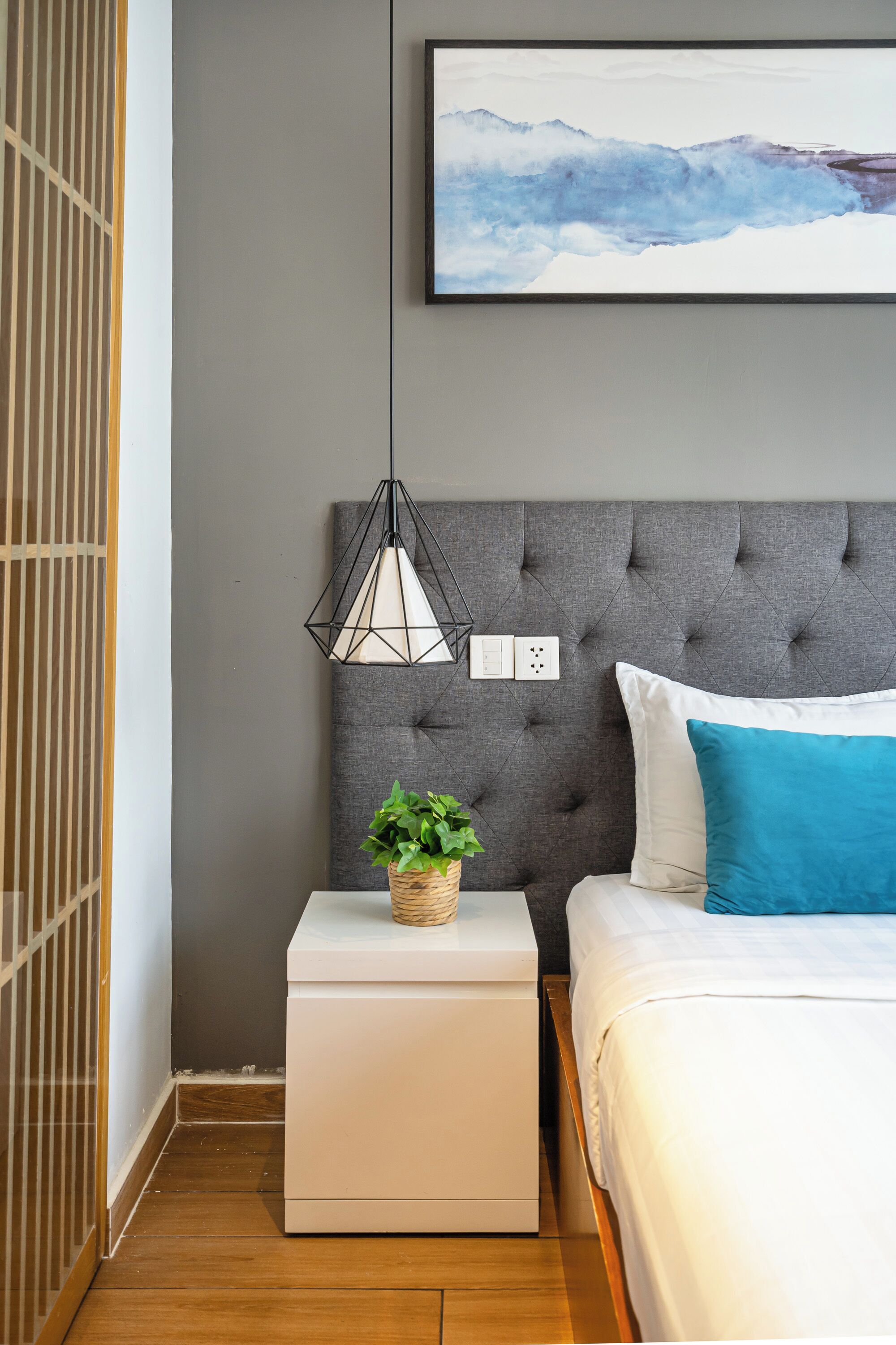A bedroom with a grey upholstered headboard, a bedside table with a white lamp, a potted plant, and a bed with white and blue bedding.